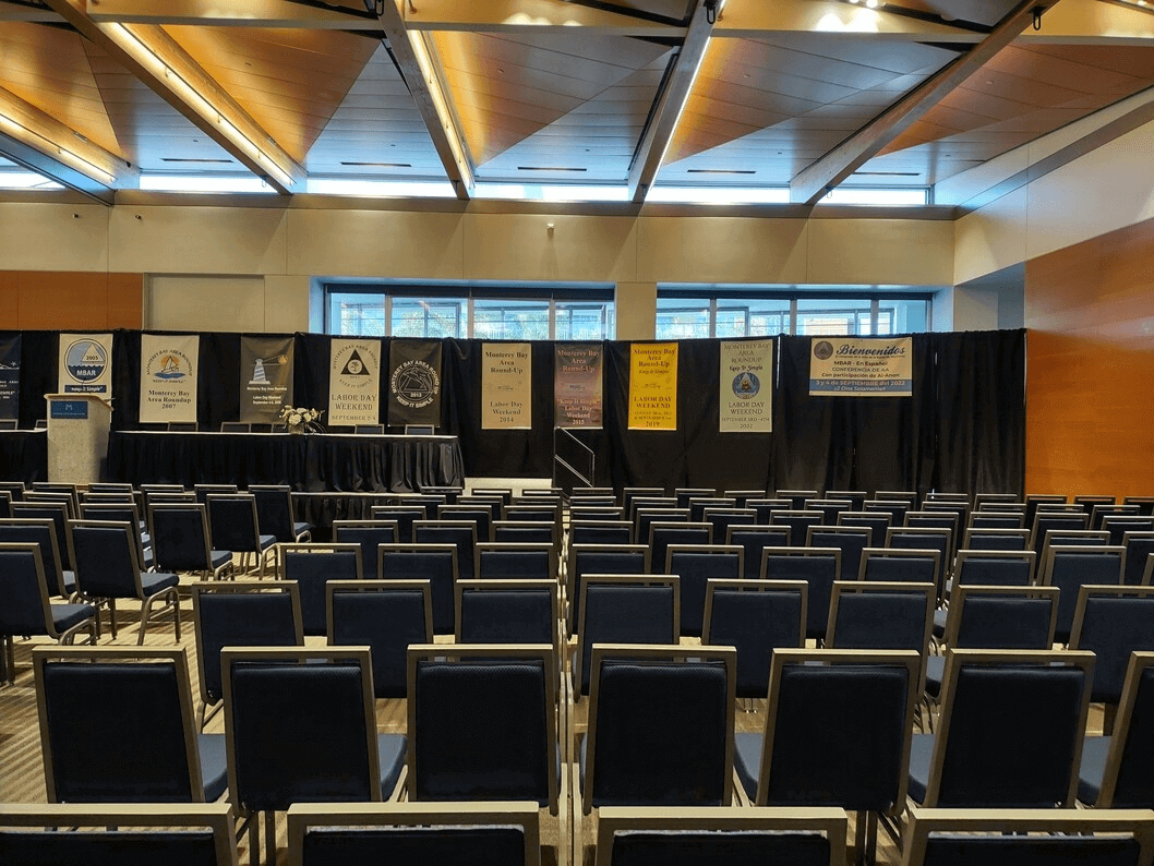 Monterey Conference Center meeting room, chairs lined up facing a stage, and past A.A. conference banners.